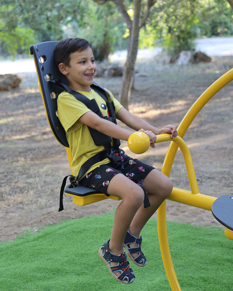 Ein kleiner Junge trägt ein Sicherheitsgeschirr und benutzt die inklusive Wippe Dennis auf einem Spielplatz.